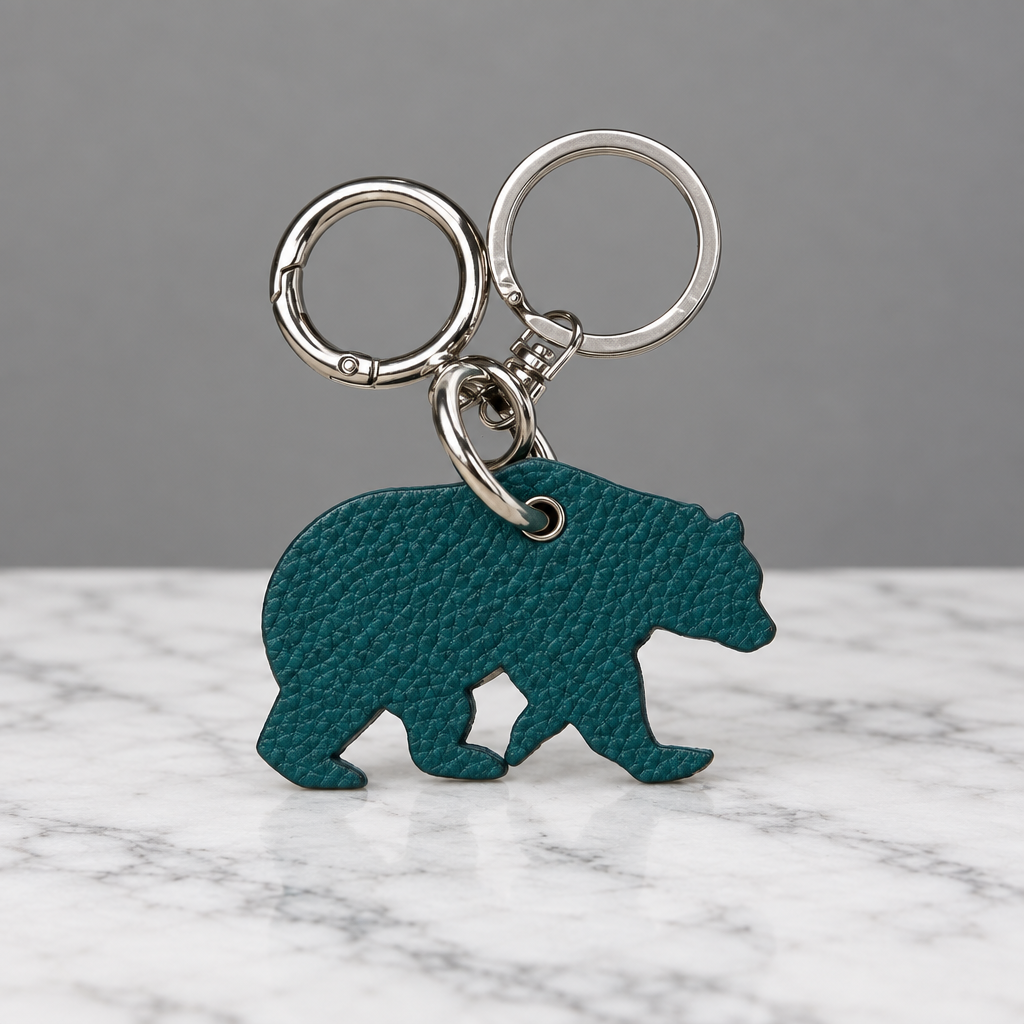 A teal bear-shaped keychain with a textured leather-like finish and silver metal key rings, shown on a light beige surface.