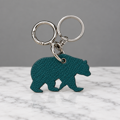 A teal bear-shaped keychain with a textured leather-like finish and silver metal key rings, shown on a light beige surface.